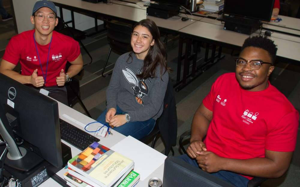 Three students are seated at a computer desk, smiling at the camera in a classroom or lab setting. Two of them, wearing matching red shirts with white logos, sit on either side of a woman in a gray sweatshirt with a hornet graphic; books and a computer.